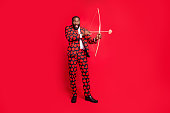 Full body photo of funny dark skin man with bow and love arrow amour cupid role see good couple wear hearts pattern suit shirt necktie tie boots outfit isolated red color background