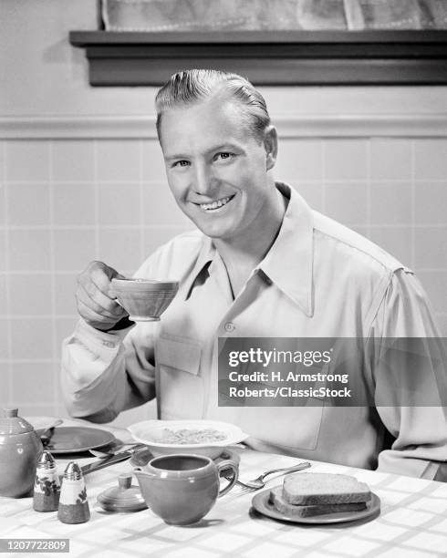 1940s smiling man looking at camera bright and early sitting eating at breakfast kitchen table holding drinking a cup of coffee.