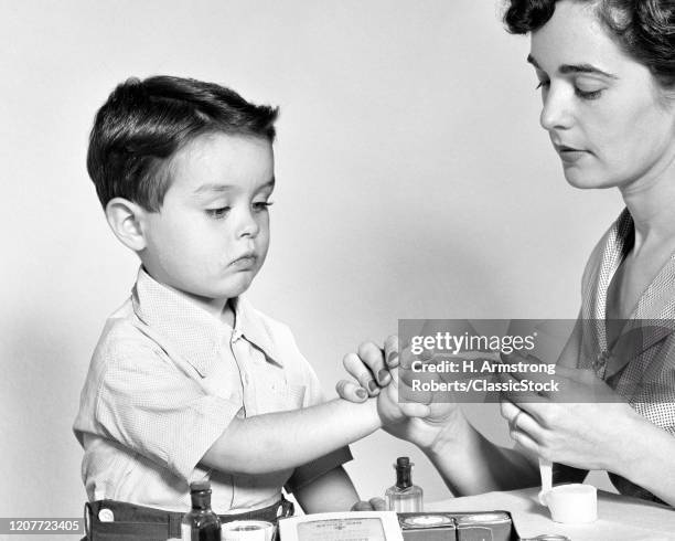 1950s mother administering first aid using rolled gauze bandage wrapping little boy's injured index finger .