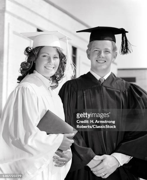 1970s portrait smiling teenage student couple in graduation robes looking at camera.
