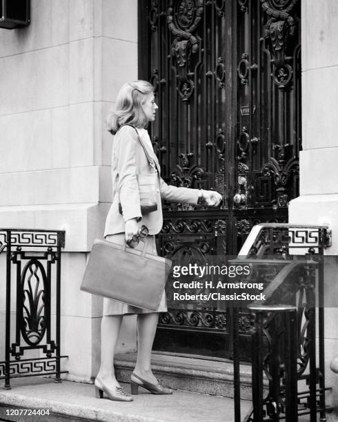 1970s dressed for success blond business woman carrying leather briefcase opening imposing large wrought iron office front door .