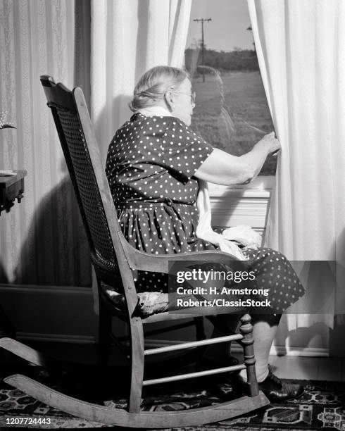 1960s senior woman sitting in rocking chair by window looking out for anticipated visitor or nosey biddy spying on neighbors.