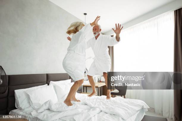 silly senior couple having fun in hotel room jumping on a bed - old couple jumping stock-fotos und bilder