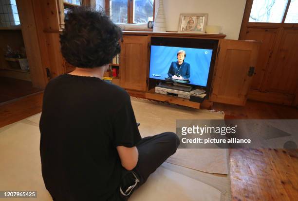 March 2020, Bavaria, Kaufbeuren: In a living room, a young man watches the broadcast of a speech by the German Chancellor Angela Merkel on the...