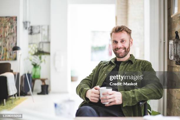 portrait of smiling male entrepreneur with cup sitting at home office - portrait home stock-fotos und bilder