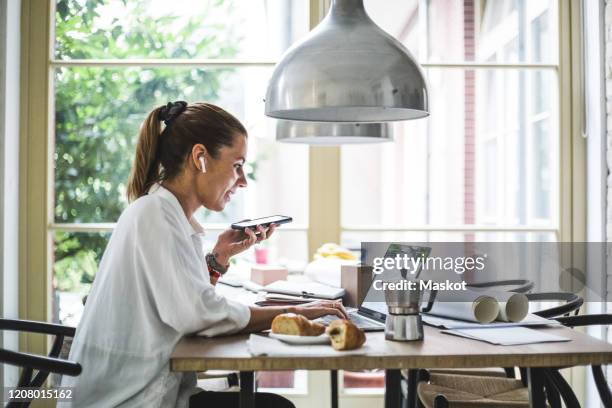 smiling female entrepreneur using laptop while talking through smart phone at home office - multitareas fotografías e imágenes de stock
