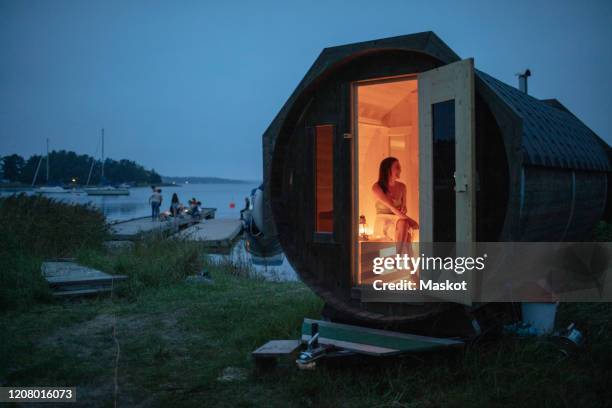 full length of mid adult woman sitting in sauna cabin by sea against sky - sauna ストックフォトと画像