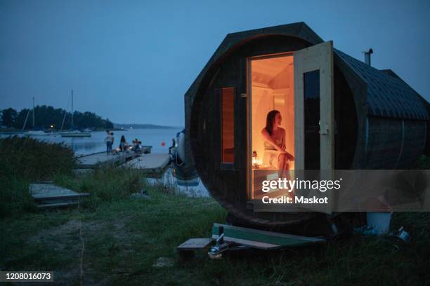 full length of mid adult woman sitting in sauna cabin by sea against sky - sauna stock pictures, royalty-free photos & images