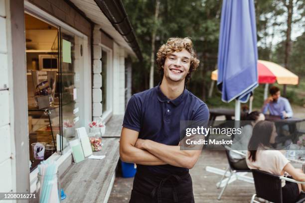 portrait of young owner with arms crossed standing by concession stand while customer sitting in background - deeltijdbaan stockfoto's en -beelden