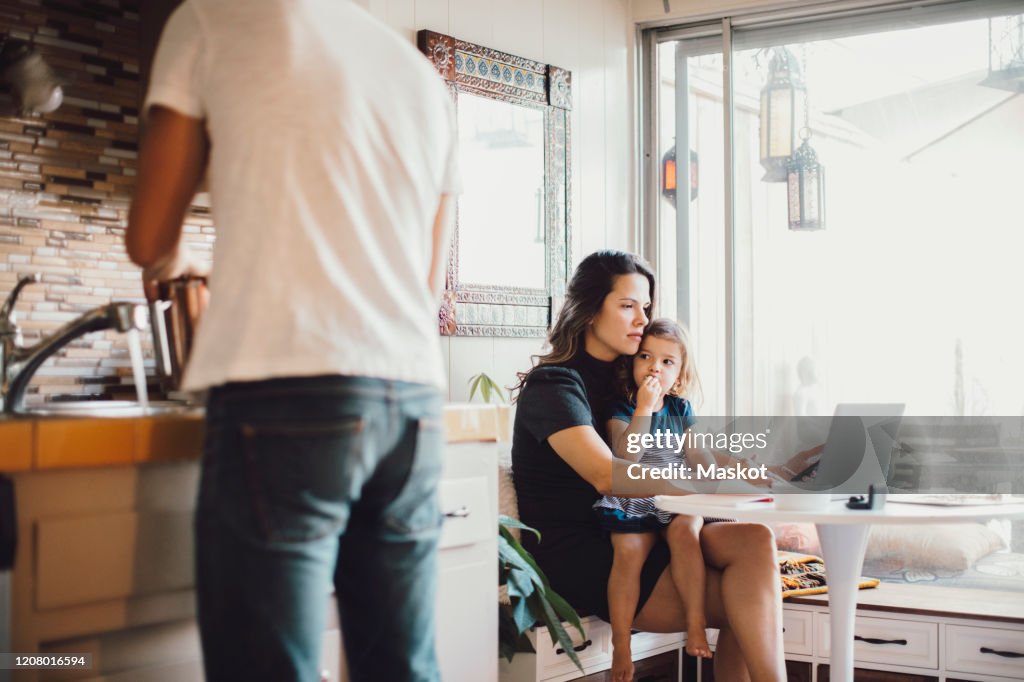 Mother working on laptop while daughter looking at father standing in kitchen