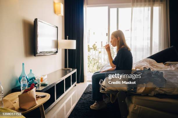 side view of girl watching television while sitting on bed in hotel room - tv room side imagens e fotografias de stock