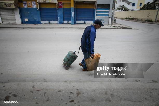 An elderly man carries a gas bottle on a cart with one hand and a bag of bread with the other hand, in Ariana downtown, northern Tunisia, at the...