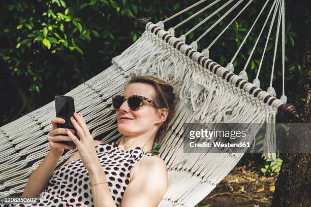 smiling woman lying in a hammock using cell phone - hammock phone stock pictures, royalty-free photos & images