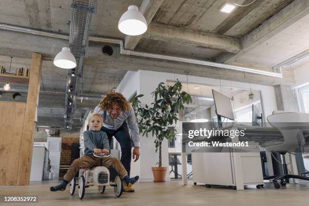 businessman pushing son on toy car in office - kid office stock-fotos und bilder