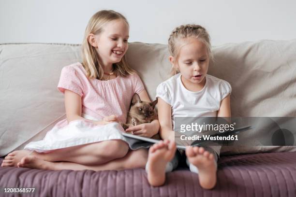 two little girls sitting barefoot on the couch with burmese cat watching a book - burmese cat stock-fotos und bilder