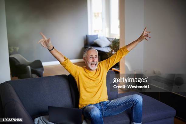 mature man sitting on couch at home cheering - cheering stock-fotos und bilder