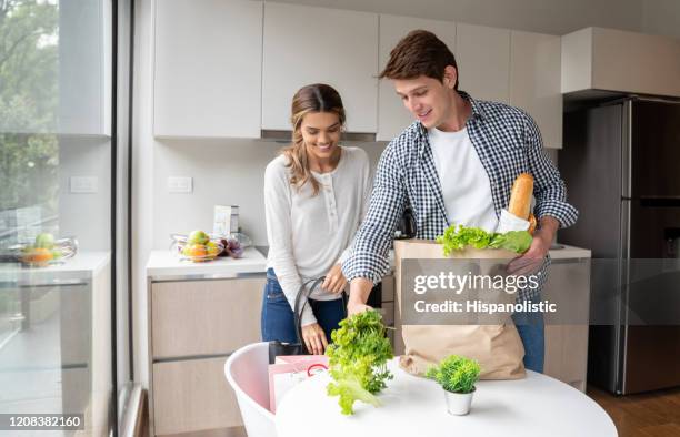 unga latinamerikanska par anländer hem med sina livsmedel uppackning medan leende - latin american and hispanic shopping bags bildbanksfoton och bilder