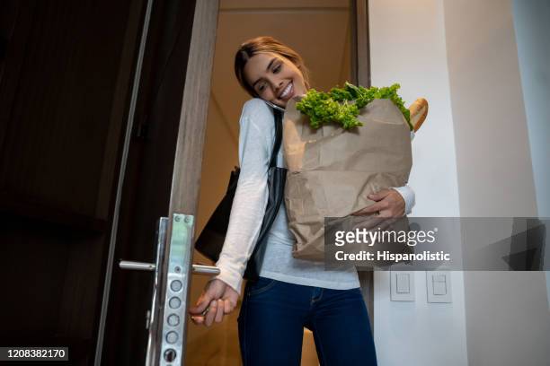 mulher bonita chegando em casa carregando mantimentos em um saco de papel abrindo a porta e em um telefonema - entrando - fotografias e filmes do acervo