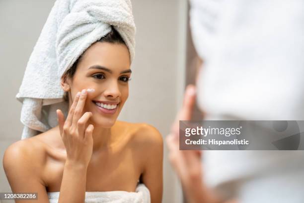 bella donna che applica crema viso dopo una doccia rilassante mentre sorride allo specchio - routine foto e immagini stock