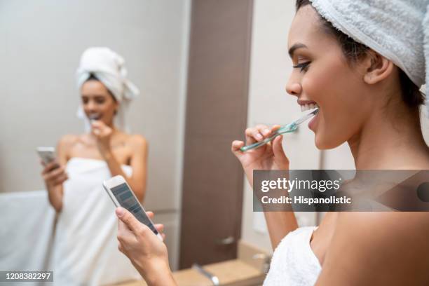 beautiful woman getting ready brussing her teeth after taking a shower texting on smartphone smiling - brush teeth phone stock pictures, royalty-free photos & images