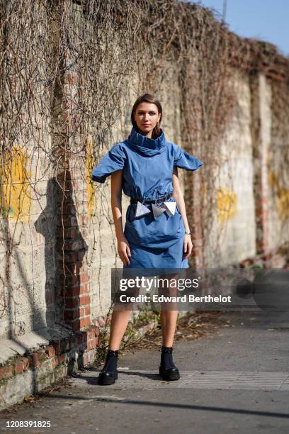 Landiana Cerciu wears a blue dress with large collar and shoulder pads, a belt with attached triangular mini bags, boots, outside BOSS, during Milan...