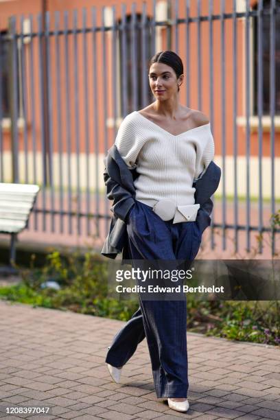 Guest wears an off-shoulder white wool pullover, a belt bag, a gray jacket, blue flared pants, white pointy shoes, outside BOSS, during Milan Fashion...