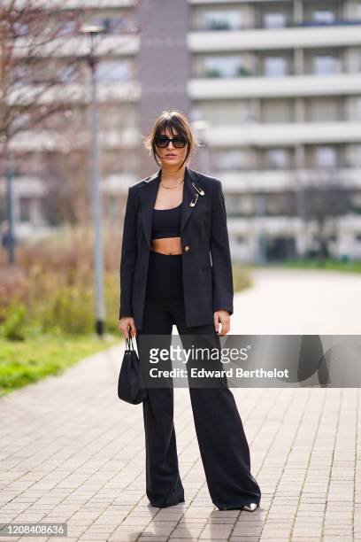 Carlotta Rubaltelli wears suglasses, a black blazer jacket with attached large safety pins, black bras, flared pants, pointy shoes, a bag, outside...