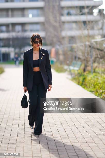 Carlotta Rubaltelli wears suglasses, a black blazer jacket with attached large safety pins, black bras, flared pants, pointy shoes, a bag, outside...