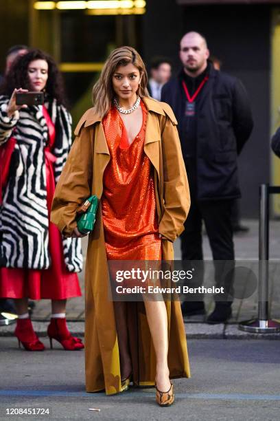 Rola wears earrings, a chain necklace, a glittering orange dress with a plunging neckline, a camel oversized trench coat, a green woven leather...