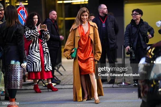 Rola wears earrings, a chain necklace, a glittering orange dress with a plunging neckline, a camel oversized trench coat, a green woven leather...