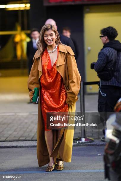 Rola wears earrings, a chain necklace, a glittering orange dress with a plunging neckline, a camel oversized trench coat, a green woven leather...