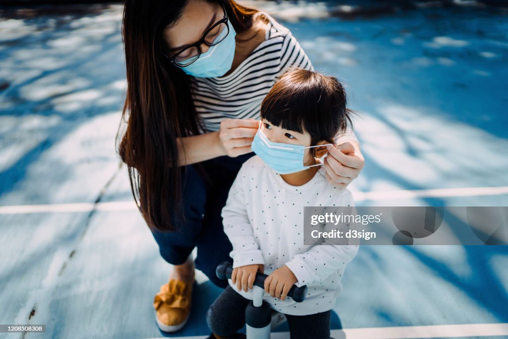 Young mother putting on surgical mask for little daughter in the playground to prevent the spread of cold and flu and viruses