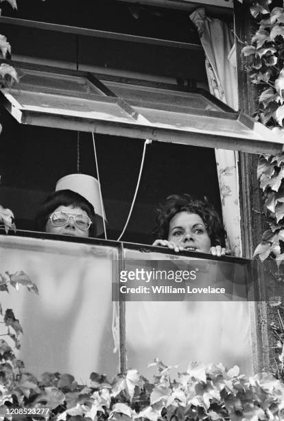 American tennis player Billie Jean King and Australian tennis player Evonne Goolagong peering out of an awning window, with their faces partially...