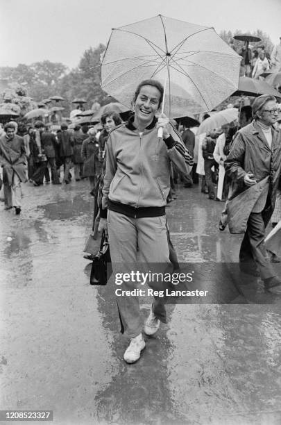 British tennis player Virginia Wade wearing a tracksuit while taking shelter under an umbrella as she walks through the crowds during the 1973 John...