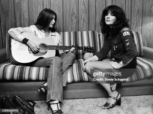 American musicians Emmylou Harris , on an acoustic guitar, and Linda Ronstadt, dressed in a Cub Scout uniform, sit together on a couch, Los Angeles,...