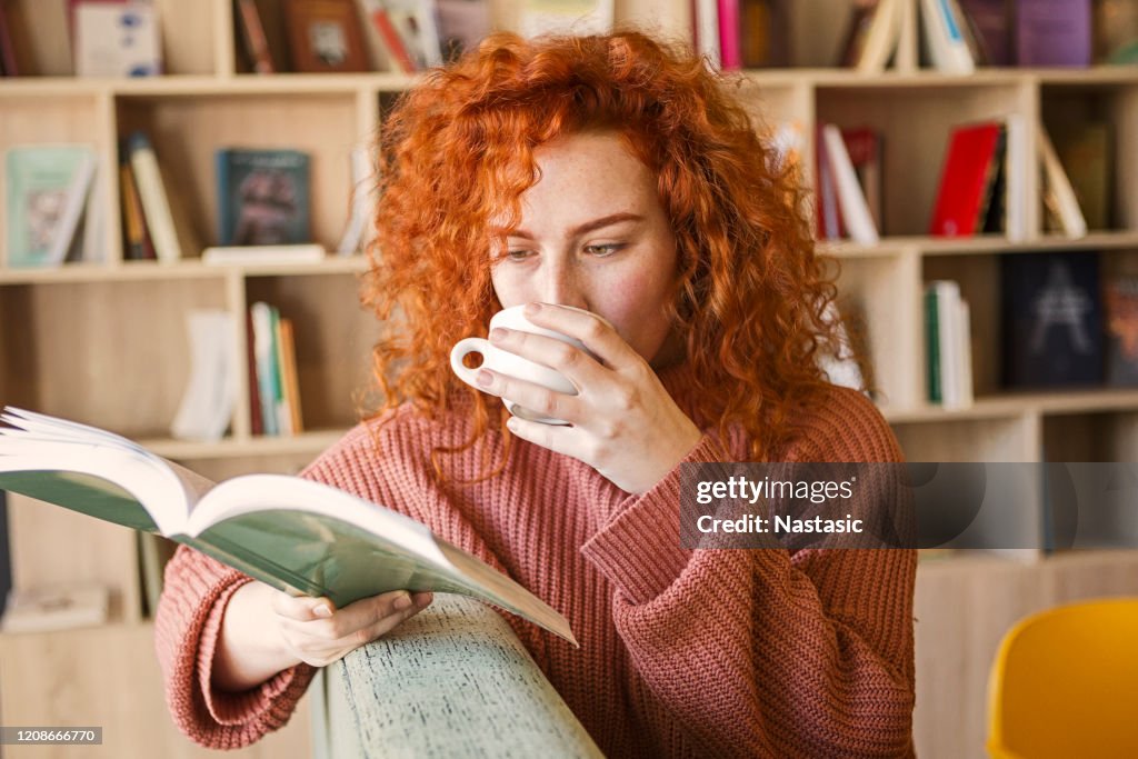 De zitting van de vrouw op bank met mok van koffie in boekhandel die een boek leest