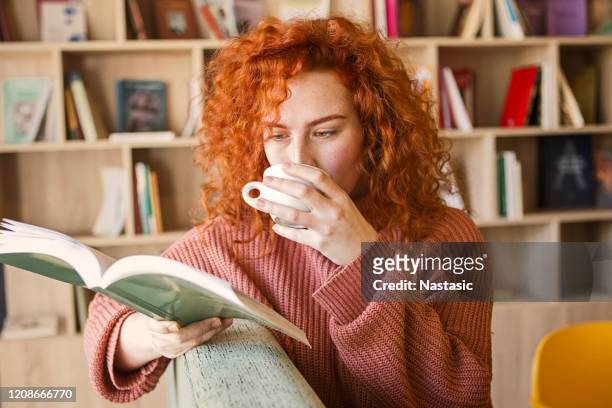 de zitting van de vrouw op bank met mok van koffie in boekhandel die een boek leest - reading stockfoto's en -beelden