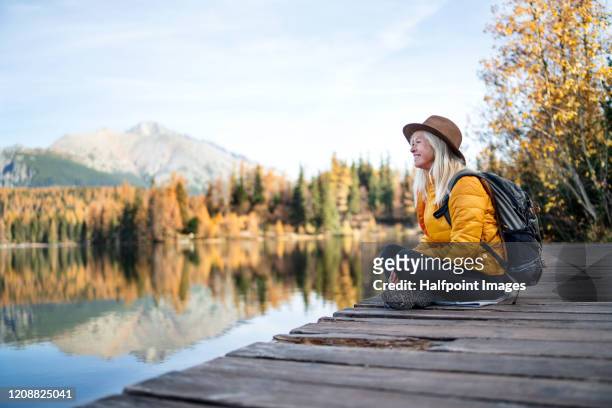 side view of senior woman hiking in autumn nature, resting by lake. - season stock pictures, royalty-free photos & images