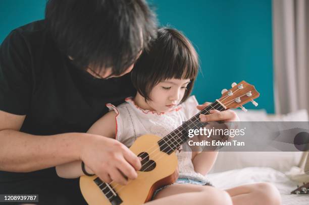 una giovane figlia cinese asiatica che impara a giocare ukulele da suo padre in camera da letto sul letto durante il fine settimana - plucking an instrument foto e immagini stock