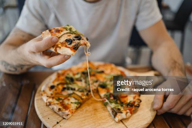 caucasian young man with hat eating a pizza with vegetables on a wooden table - lunch cheese foto e immagini stock