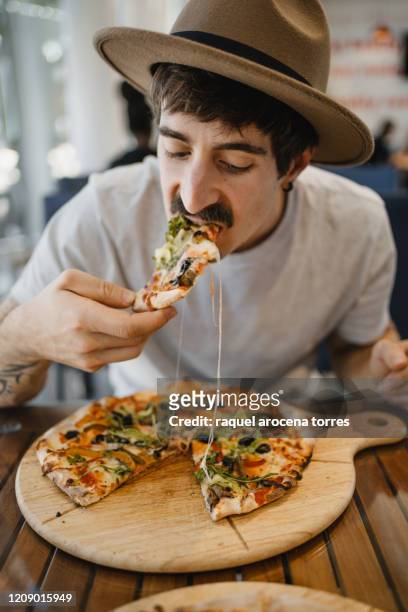 caucasian young man with hat eating a pizza with vegetables on a wooden table - pizzeria bildbanksfoton och bilder