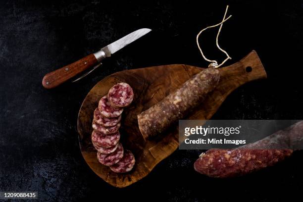 high angle close up of knife, sliced salami on wooden cutting board on black background. - salami stock pictures, royalty-free photos & images