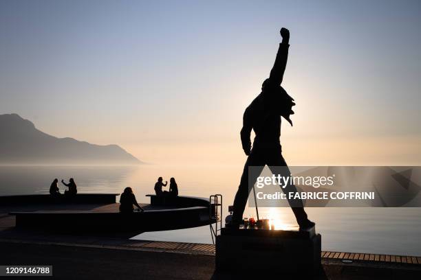 People sit on a bench at sunset close to a statue of the rock band Queen's late singer Freddie Mercury, in silhouette, erected on the shores of Lake...