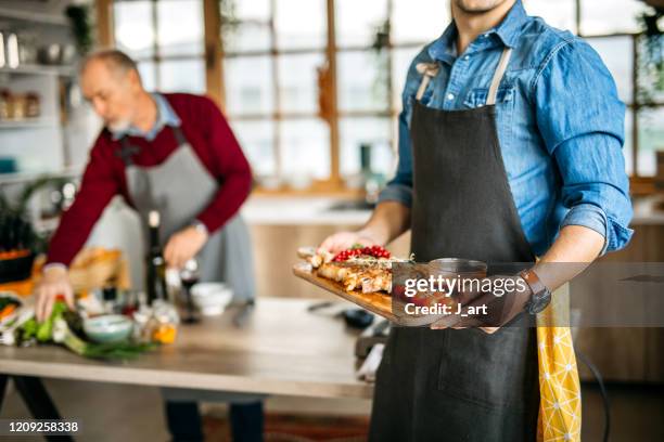 millennial generation son holding a cutting board with served meat dish and vegetables. - apron stockfoto's en -beelden