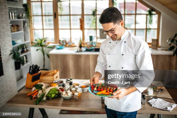 young chef holding a plate with beautifully served meat dish and vegetables. - personal chef stock pictures, royalty-free photos & images