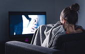 Scary horror movie on tv. Scared woman watching stream service hiding under blanket on couch at night. Sleepless person streaming series or film on television.