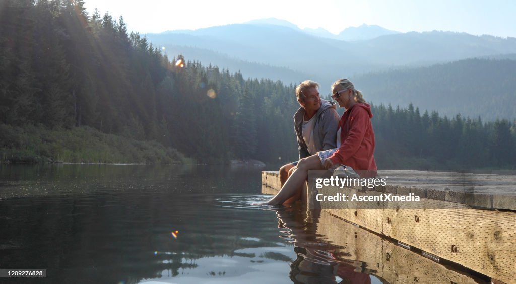 Mature couple relax on wooden pier, looks out across lake