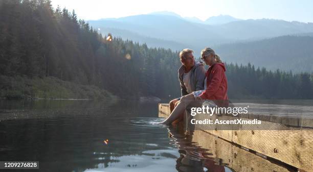 mature couple relax on wooden pier, looks out across lake - woman pier stock pictures, royalty-free photos & images