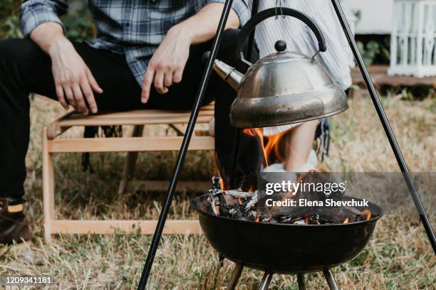 a man and a woman at a camping are watching a kettle boiling over an open fire. - tripod ストックフォトと画像