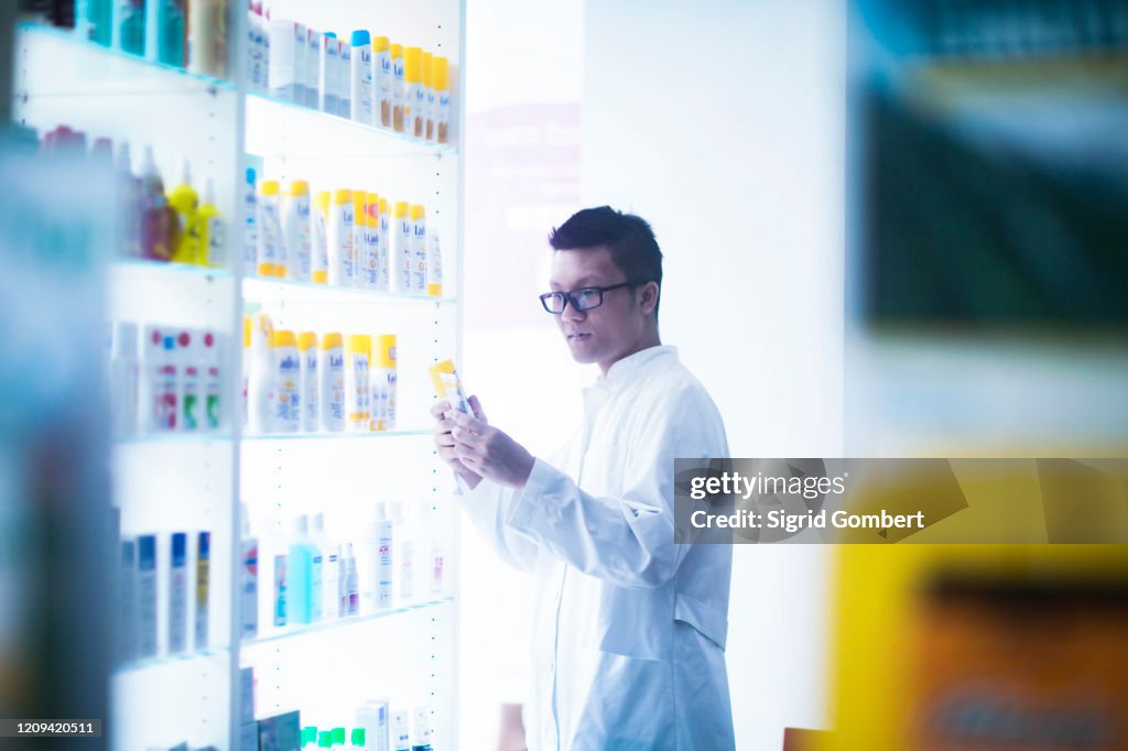 Male Asian pharmacist wearing glasses standing next to shelves with suntan lotions in pharmacy.